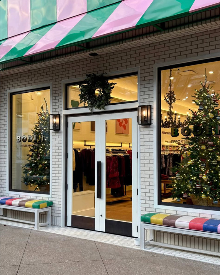 A storefront with white brick walls, a green and pink striped awning, double glass doors, large display windows, and colorful bench seating. Christmas trees and wreaths decorate the entrance.
