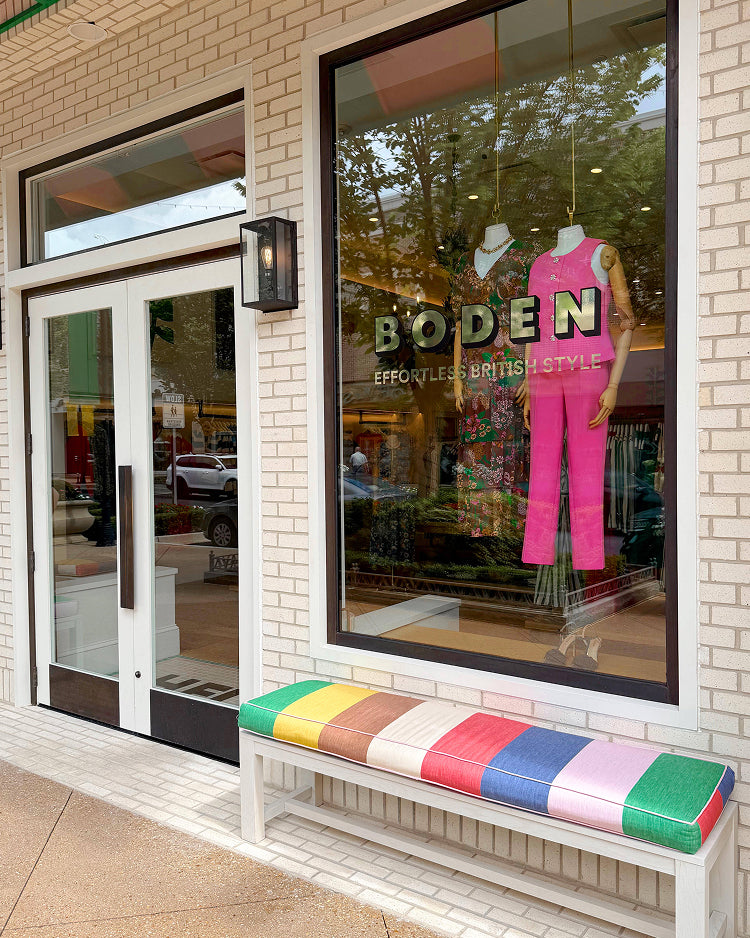 A storefront window display for BODEN Effortless British Style clothing, featuring a mannequin in a bright pink two-piece outfit. Below the window is a white bench with a colorful striped cushion in green, blue, pink, yellow, brown, and white.