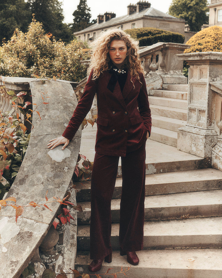 standing on stone steps in a formal garden setting, wearing a deep burgundy double-breasted velvet blazer with matching wide-leg trousers and platform shoes. A jeweled collar detail adds embellishment to the tailored look, while the historic stone balustrades and autumn foliage create an elegant heritage-inspired backdrop.