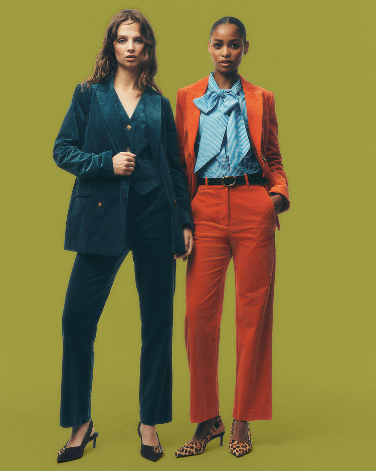 Two models posing in matching tailored suits against a solid olive green background. The model on the left wears a dark teal velvet pantsuit, and the model on the right wears a vibrant orange blazer with a light blue bow tie shirt and orange trousers.
