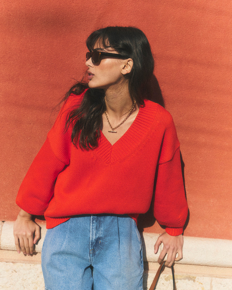 Woman wearing a red sweater and blue jeans against an orange wall.