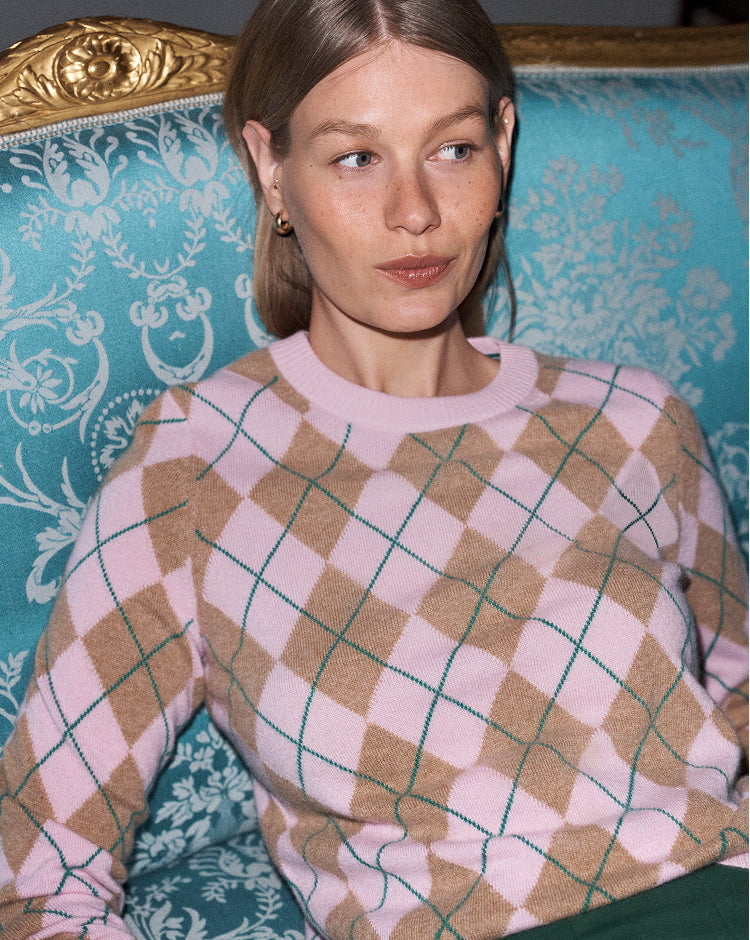 A woman wearing a pink and tan argyle sweater with thin green grid lines, seated on an ornate turquoise patterned armchair with gold detailing.