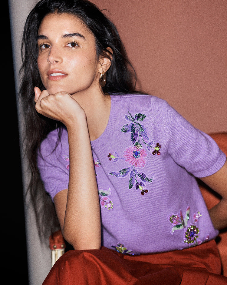A woman wearing a lavender purple short-sleeve knit sweater with colorful floral embroidery and sequins, paired with a rust-colored skirt, posing against a warm terracotta background.