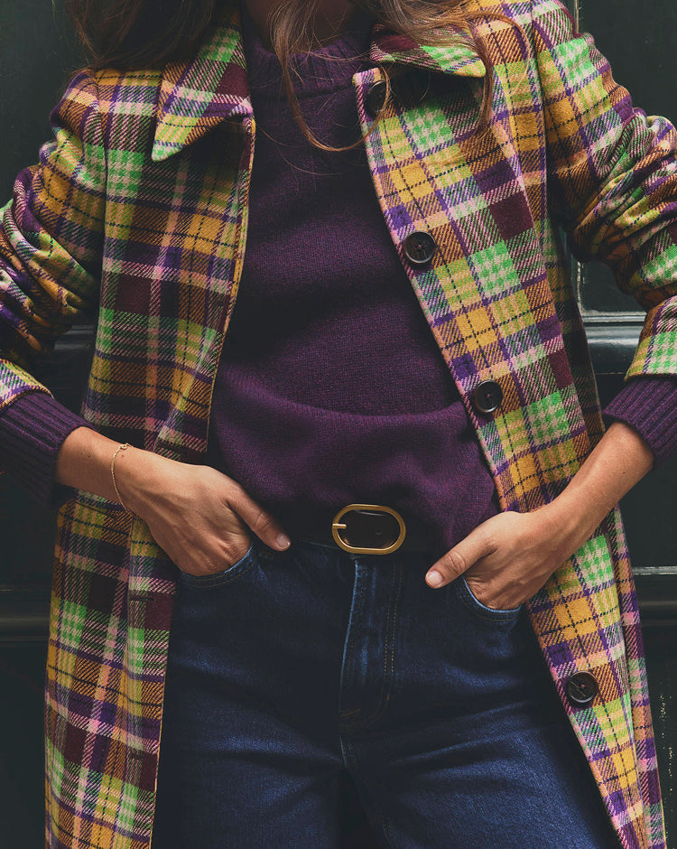 Close-up of a person wearing a purple knit sweater layered under a colourful plaid coat in shades of yellow, green and purple. The outfit is paired with dark denim jeans and a black belt with a gold oval buckle, with both hands tucked into the front pockets