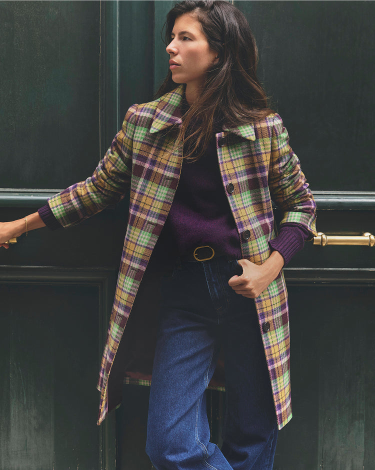 Close-up of a person wearing a purple knit sweater layered under a colourful plaid coat in shades of yellow, green and purple. The outfit is paired with dark denim jeans and a black belt with a gold oval buckle, with both hands tucked into the front pockets.