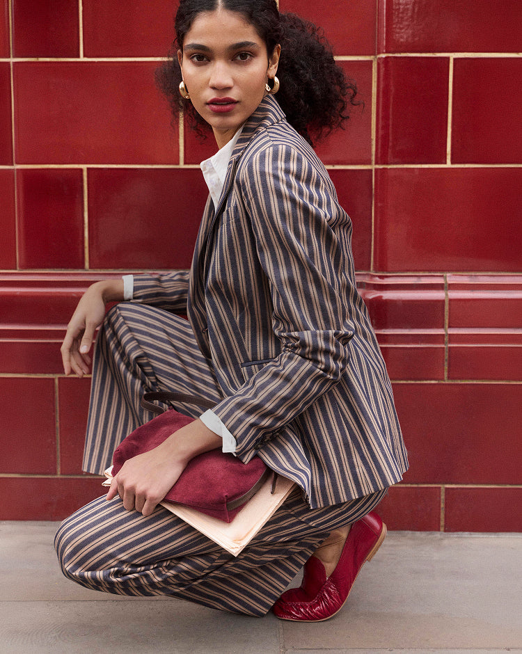 A model crouches in front of a red tiled wall wearing a brown and beige pinstripe blazer and matching trousers, holding a burgundy clutch and a light-colored book or folder.