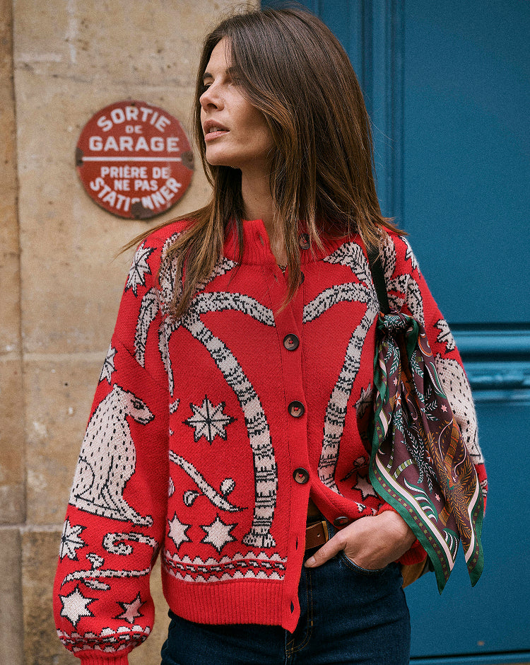 A woman wearing a vibrant red knit cardigan with a white and black tropical and animal pattern, styled with a patterned scarf and dark jeans, standing in front of a blue door and stone wall.