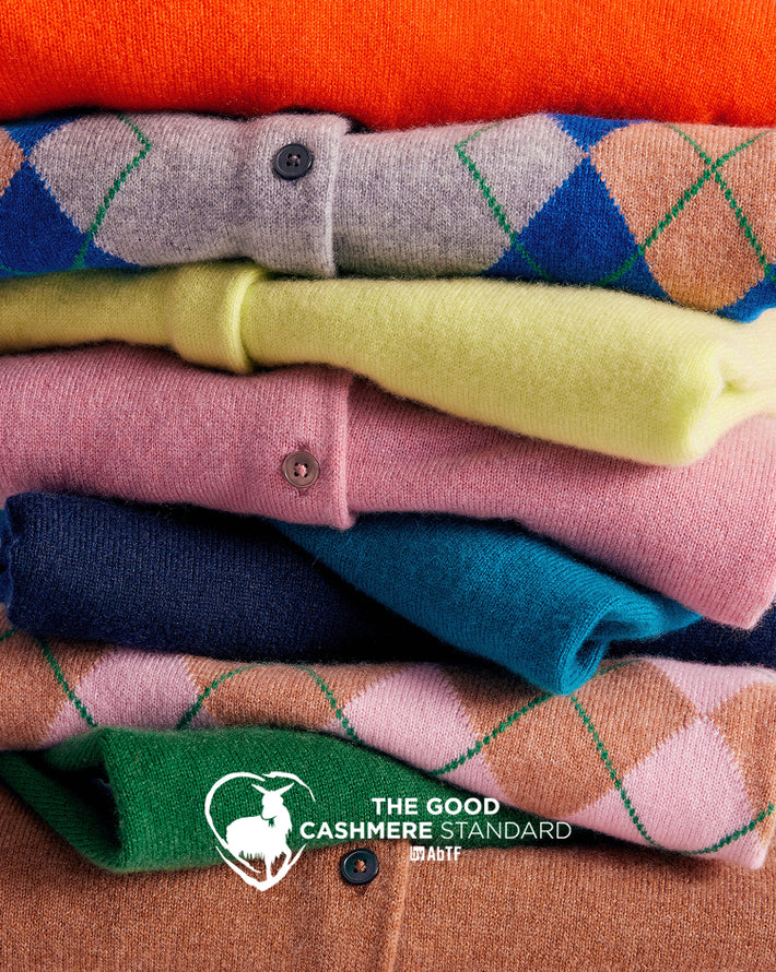 A close-up stack of folded cashmere sweaters in multiple colors, including orange, blue, yellow, pink, navy, green, and brown, with a white 'The Good Cashmere Standard' logo at the bottom.