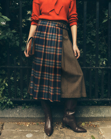 A person wearing a bright orange-red long-sleeve top, a pleated skirt with a navy, orange, and brown plaid panel and a brown textured side panel, paired with dark brown knee-high boots, standing outdoors in front of a black metal fence and greenery.