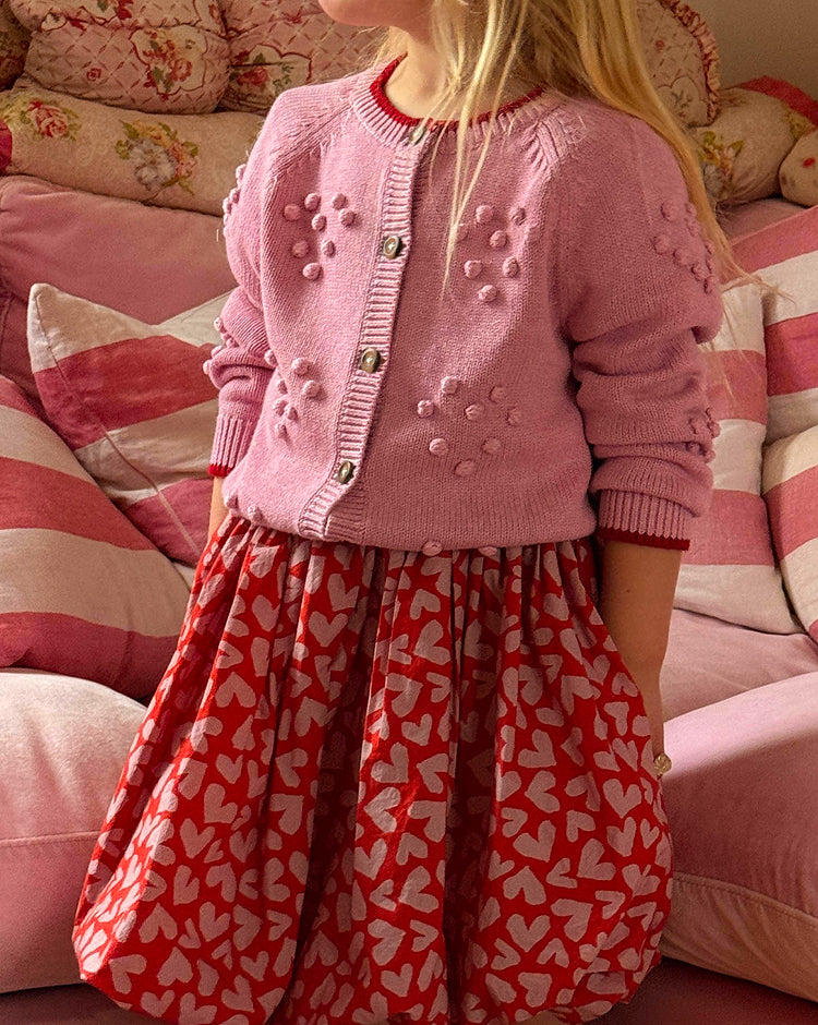 Child wearing a pink cardigan and red dress with heart patterns in a cozy room.