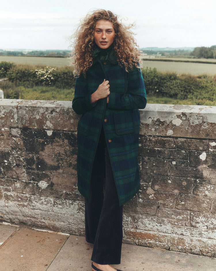 A woman with curly hair stands outdoors against a weathered stone wall, wearing a long dark green and black plaid coat over a black outfit.