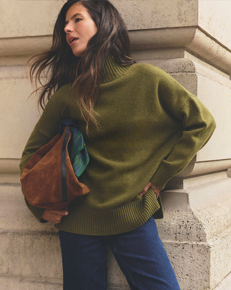 Person leaning against a stone building, wearing an oversized olive-green turtleneck sweater and dark blue jeans, while carrying a brown suede tote bag with a green tartan scarf tucked inside. The outfit is set against a neutral architectural backdrop, creating a relaxed, heritage-inspired look.