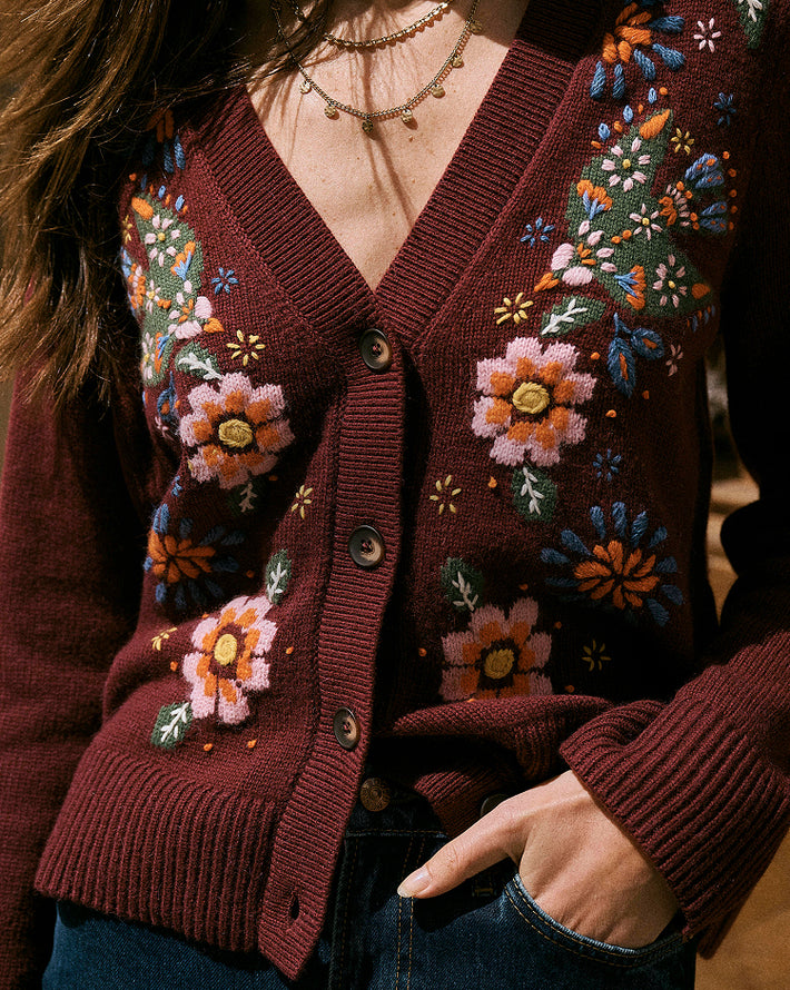 A close-up of a woman wearing a burgundy cardigan sweater with colorful floral embroidery and button-front closure, paired with blue jeans and layered gold necklaces.