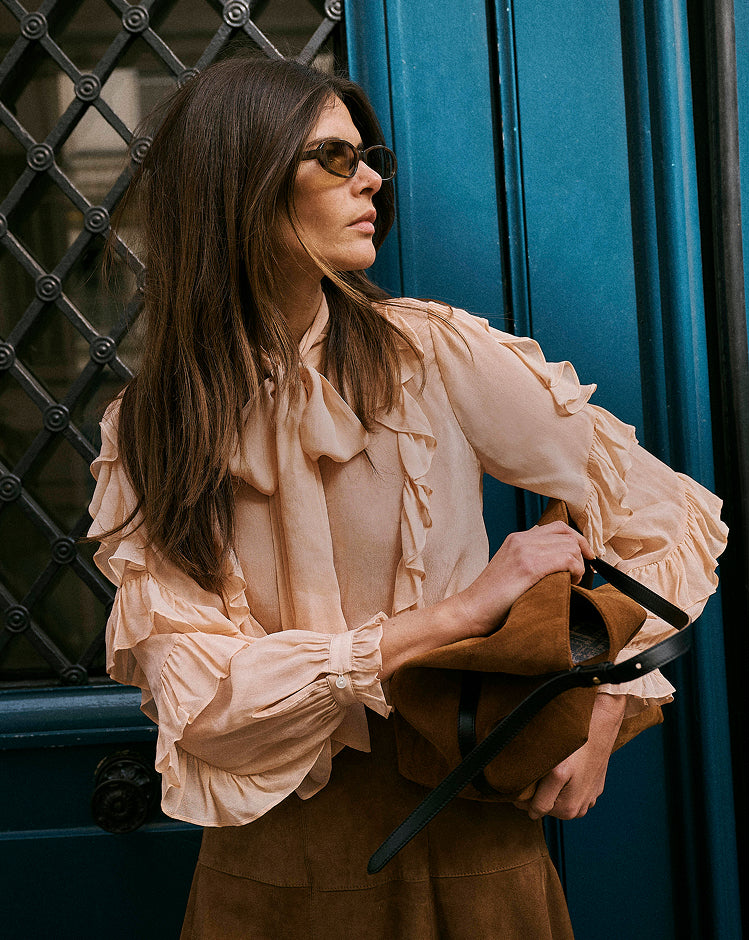 A woman wearing a beige ruffled blouse and brown skirt holds a brown leather handbag with black straps, standing in front of a dark blue door.