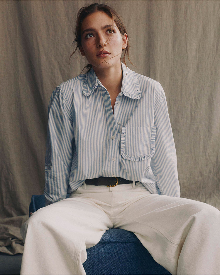 Woman wearing a light blue striped shirt and white pants against a neutral background