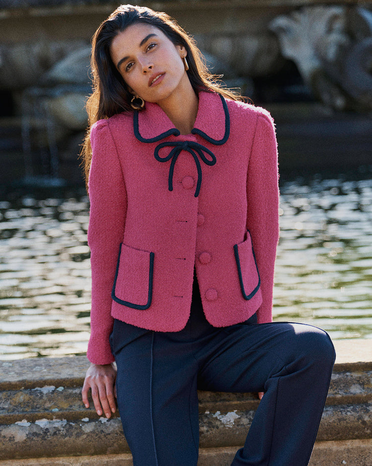 A woman wearing a pink textured jacket with black trim and a bow at the collar, paired with dark trousers, sitting outdoors near a stone fountain and water.