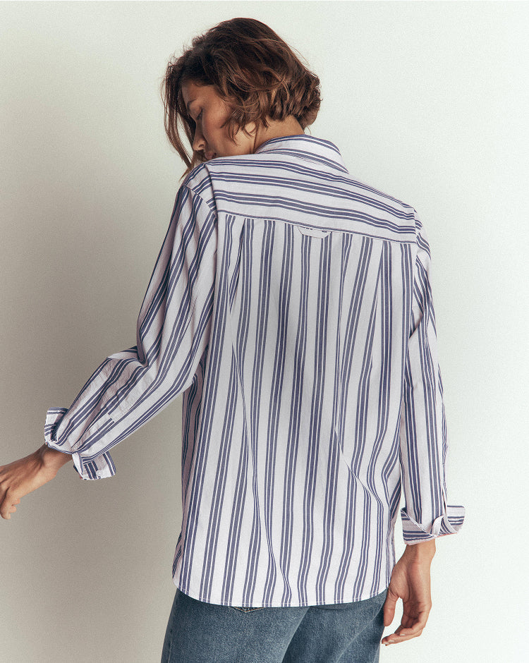 A woman shown from the back wearing a white button-up shirt with vertical navy blue stripes and blue jeans, set against a plain light gray studio background.