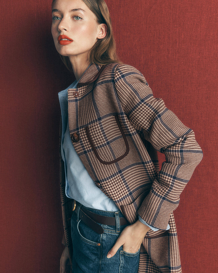 A woman wearing a beige and navy plaid blazer over a light blue shirt and dark wash jeans, posing against a textured dark red wall.