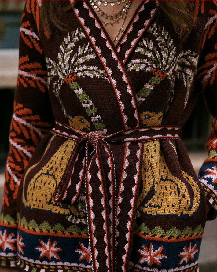 A close-up of a person wearing a dark brown knit wrap robe with a matching tie belt. The robe features a detailed multicolor pattern with green palm trees, yellow animals, and geometric borders in orange, cream, and navy blue.