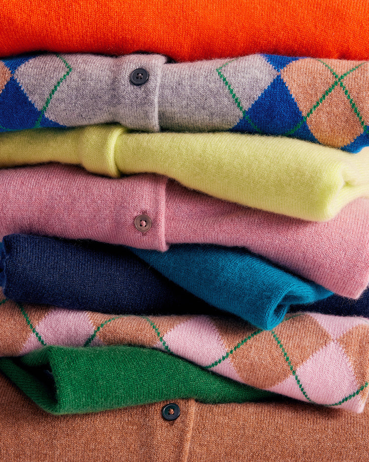 A close-up stack of folded knit cardigans in multiple colors, including orange, blue, gray, yellow, pink, navy, teal, green, and brown, with argyle and solid patterns visible.