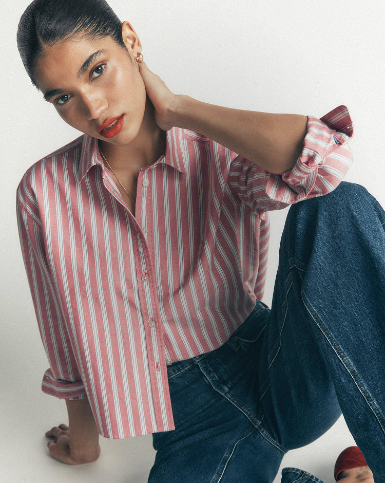 A woman wearing a pink and white vertically striped button-down shirt with rolled-up sleeves and dark blue jeans, posing against a light gray studio background.
