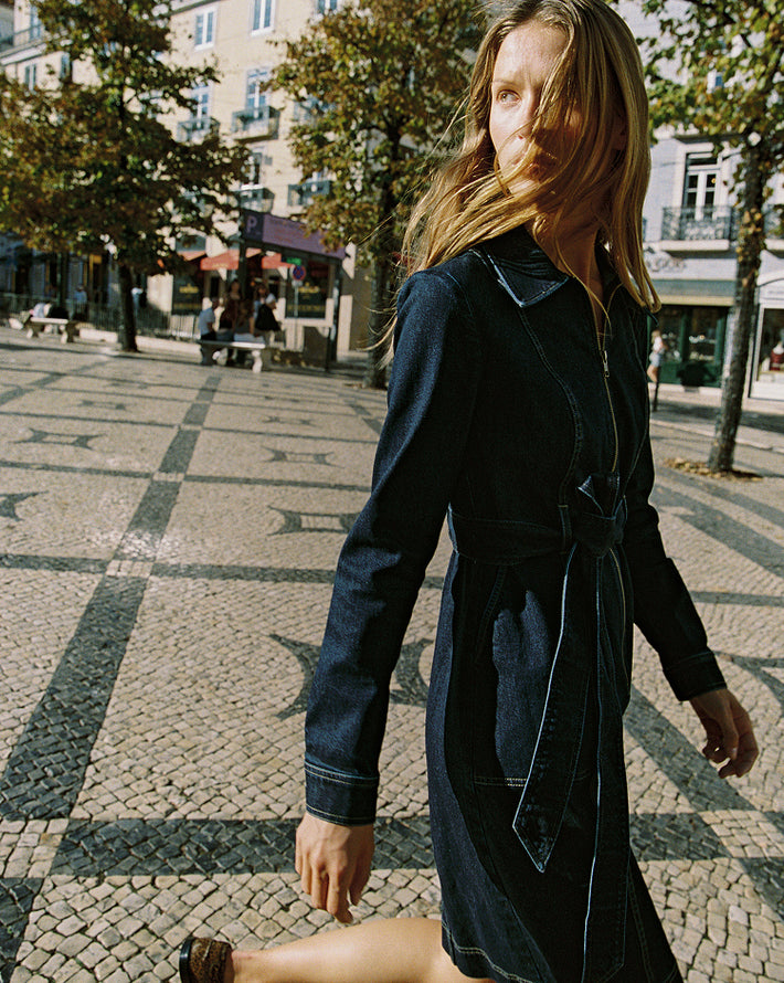 Woman in denim dress walking on a paved street with trees and buildings in the background