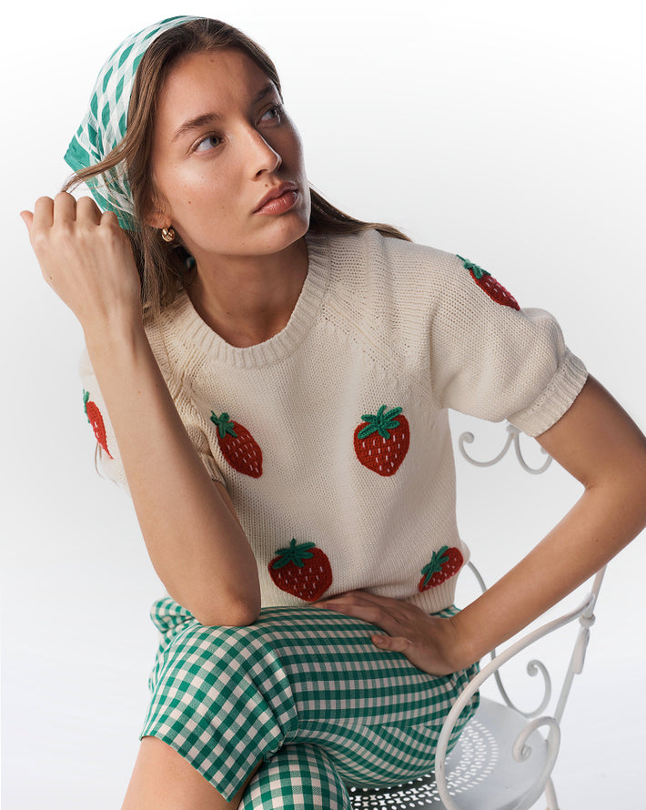 Woman wearing a cream sweater with strawberry patterns and a green checkered skirt on a white background