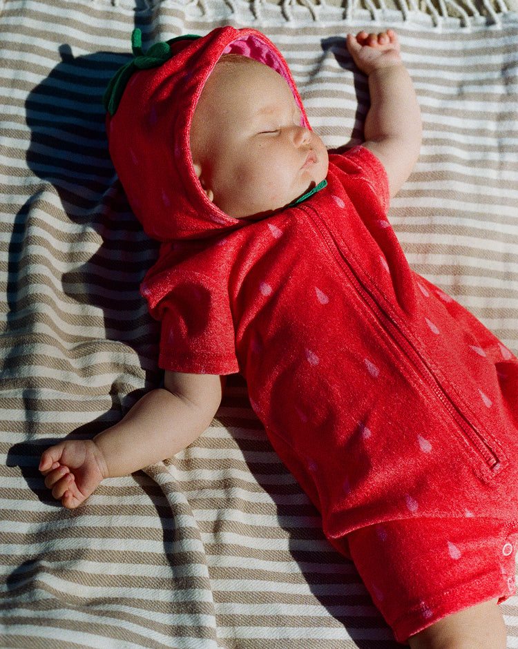 Baby wearing a red outfit with a hood, lying on a striped blanket.