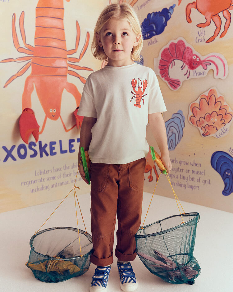 Child wearing a white t-shirt with a red lobster design, standing in front of a wall with illustrations of sea creatures.