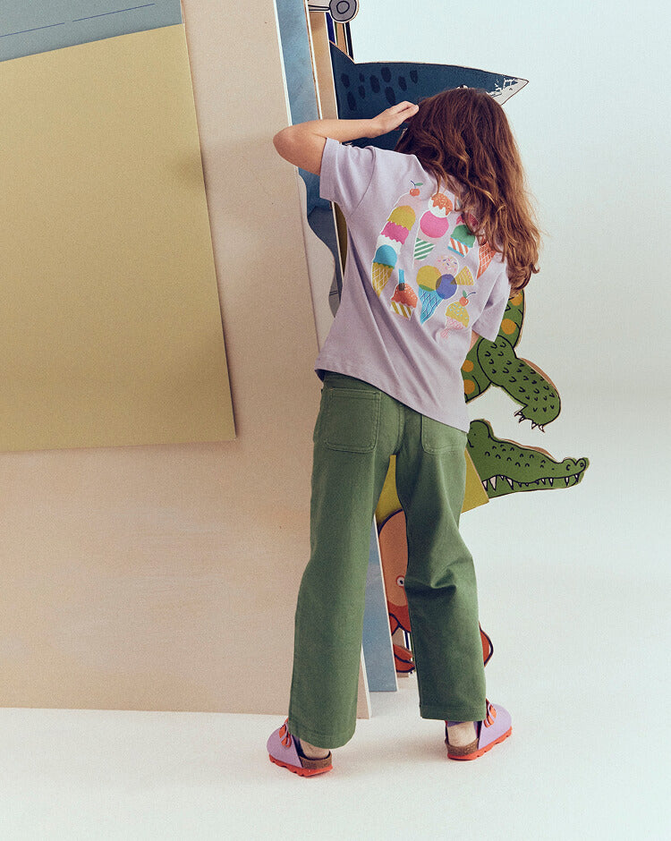 Child wearing a dinosaur-themed shirt and green pants, standing in front of a colorful wall.