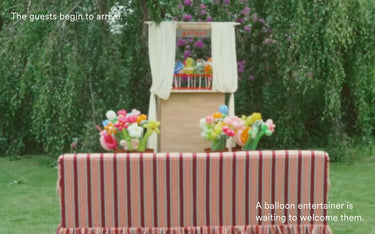 Outdoor event setup with a striped table in front of a balloon artist's chair, floral arrangements, and green foliage. Text on the image reads 'The guests begin to arrive.