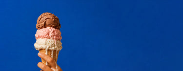 A hand holding a waffle cone filled with three scoops of ice cream in brown, pink, and white, set against a solid blue background.