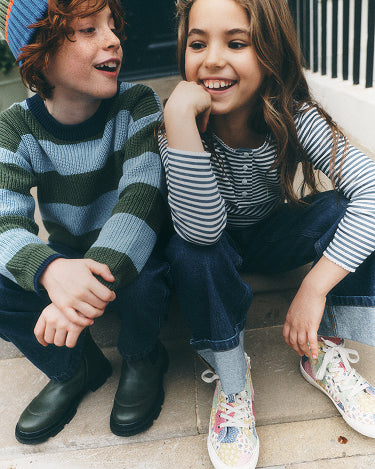Two children sitting together on outdoor steps, wearing striped knit sweaters and casual jeans. The boy wears a blue and green striped sweater with dark boots, and the girl wears a blue and white striped long-sleeve top with multicolored sneakers.