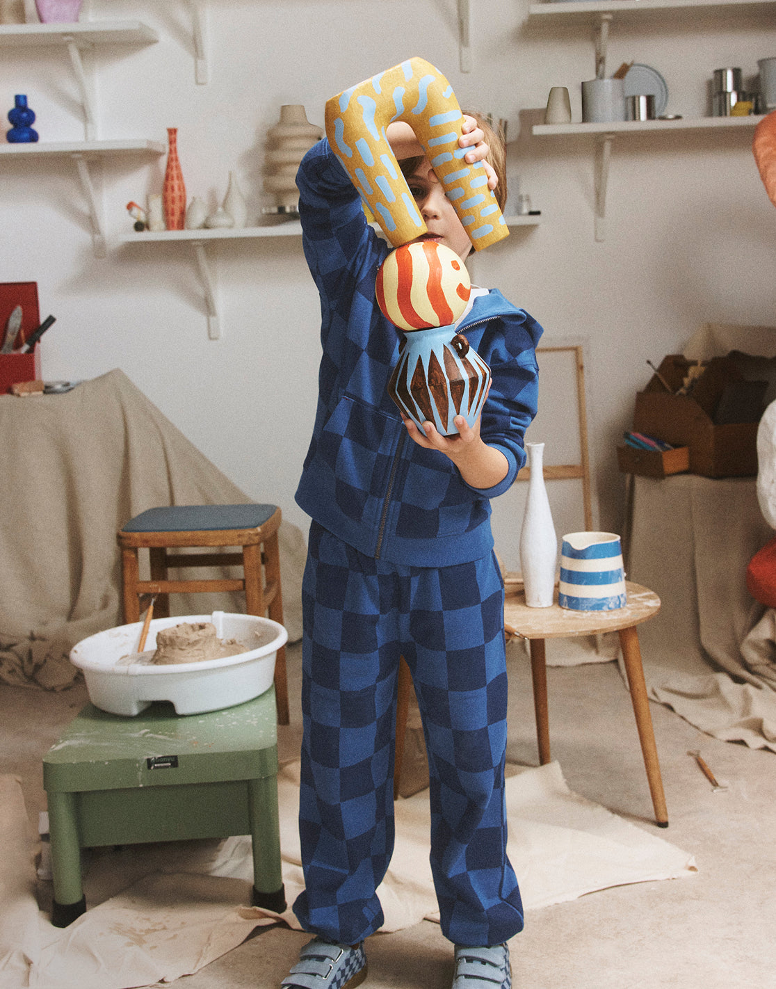 Child in a blue checkered outfit holding colorful clay sculptures in a pottery studio.