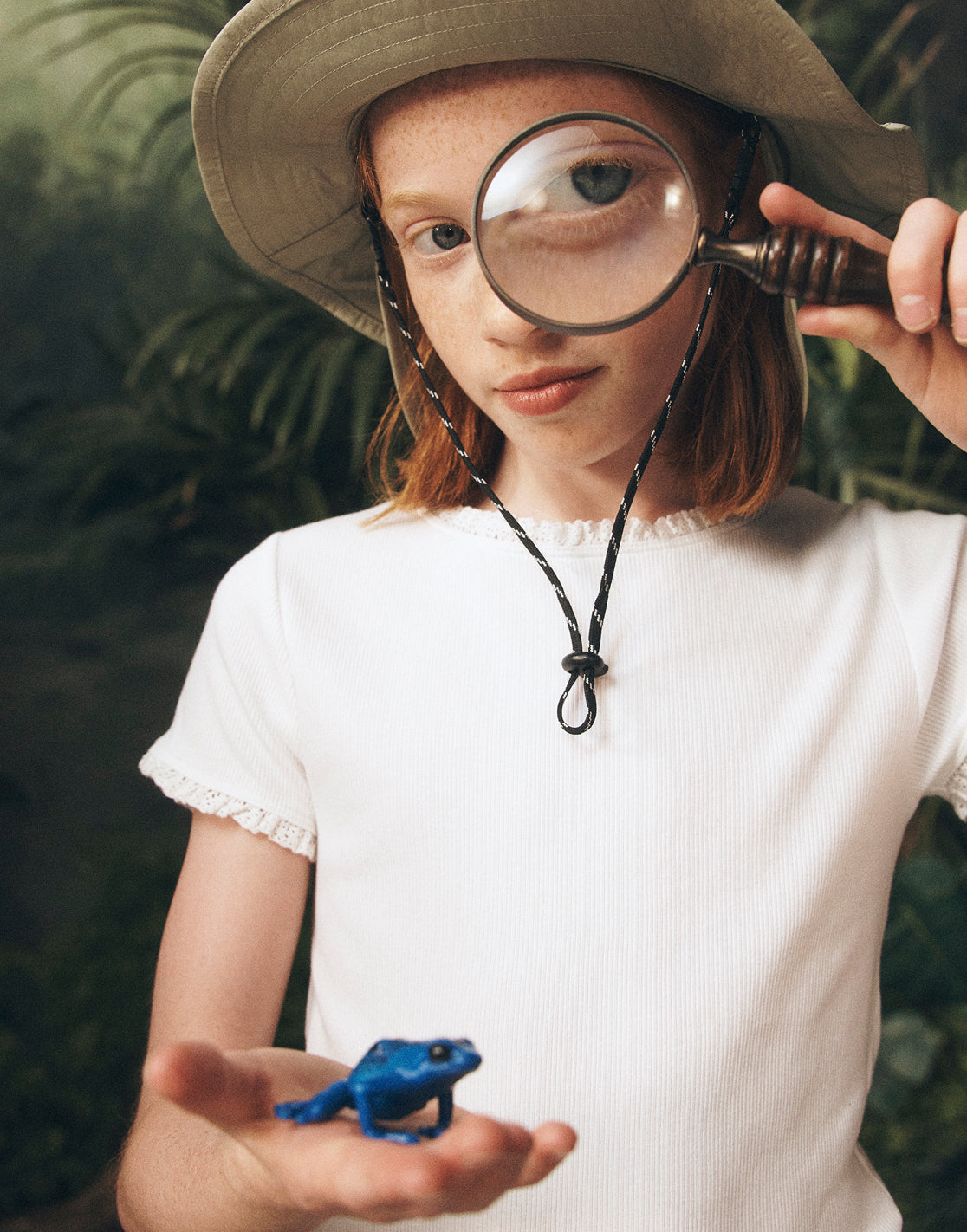 Child holding a magnifying glass over a toy frog with a natural background