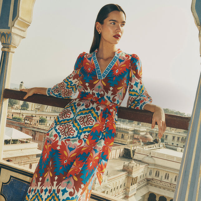 Woman in a colorful patterned dress standing on a balcony with a cityscape view.