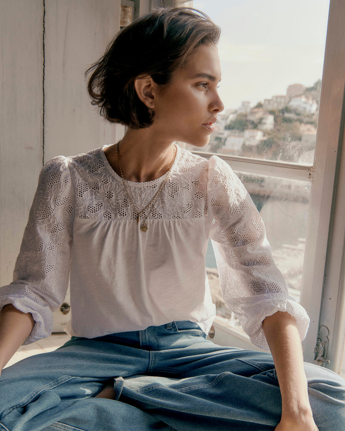 Woman sitting by a window wearing a white lace top and blue jeans, looking out.