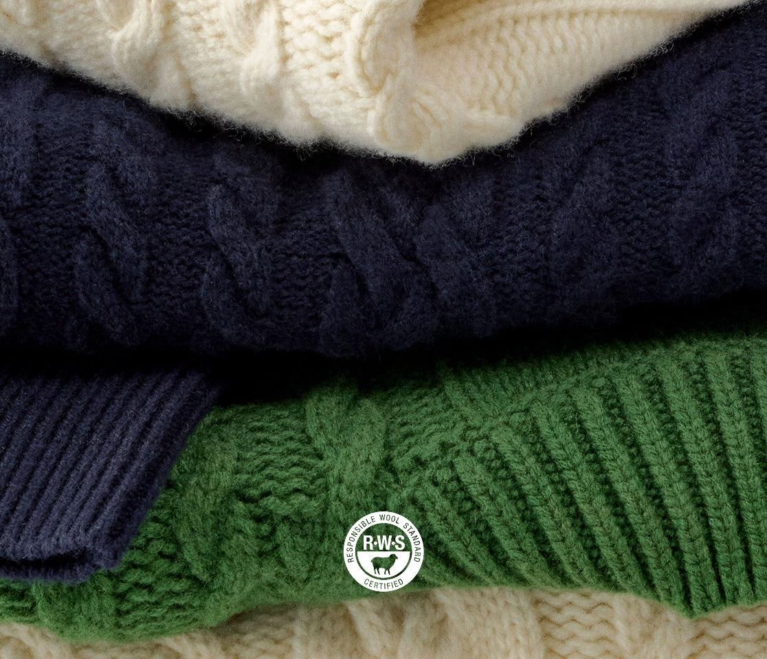 Stack of folded cable knit sweaters in navy, green, and cream with a visible brand logo.