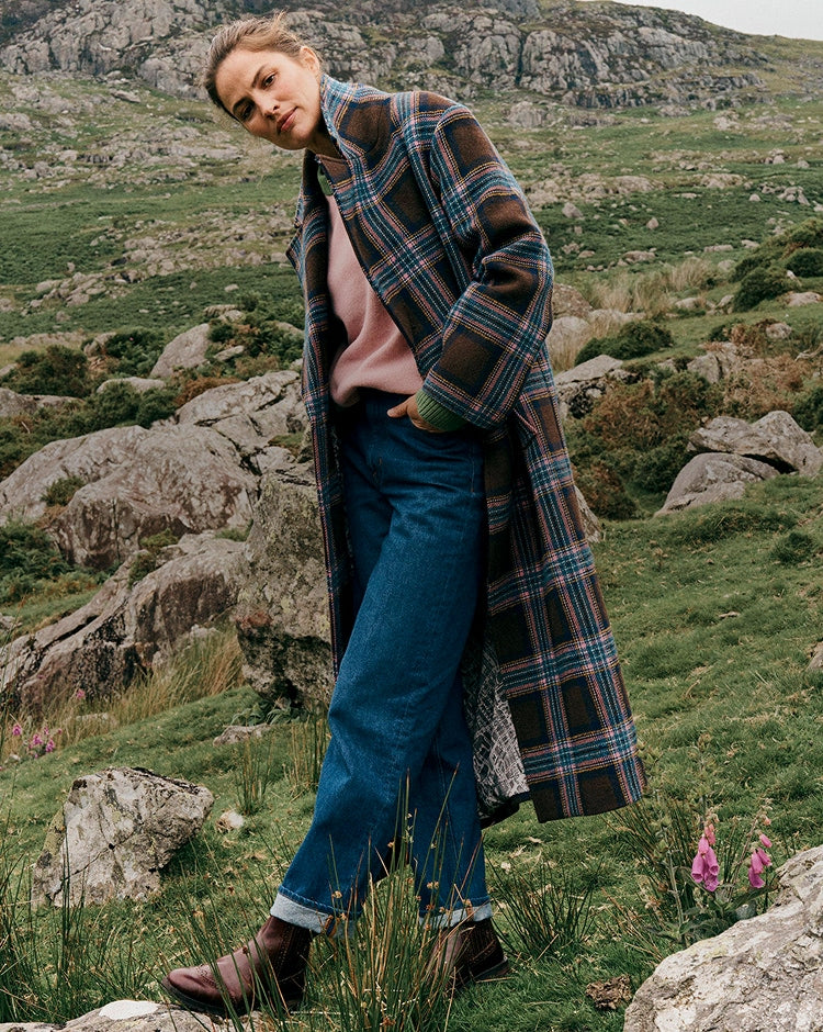 Person wearing a plaid coat and blue jeans standing in a rocky, grassy landscape.