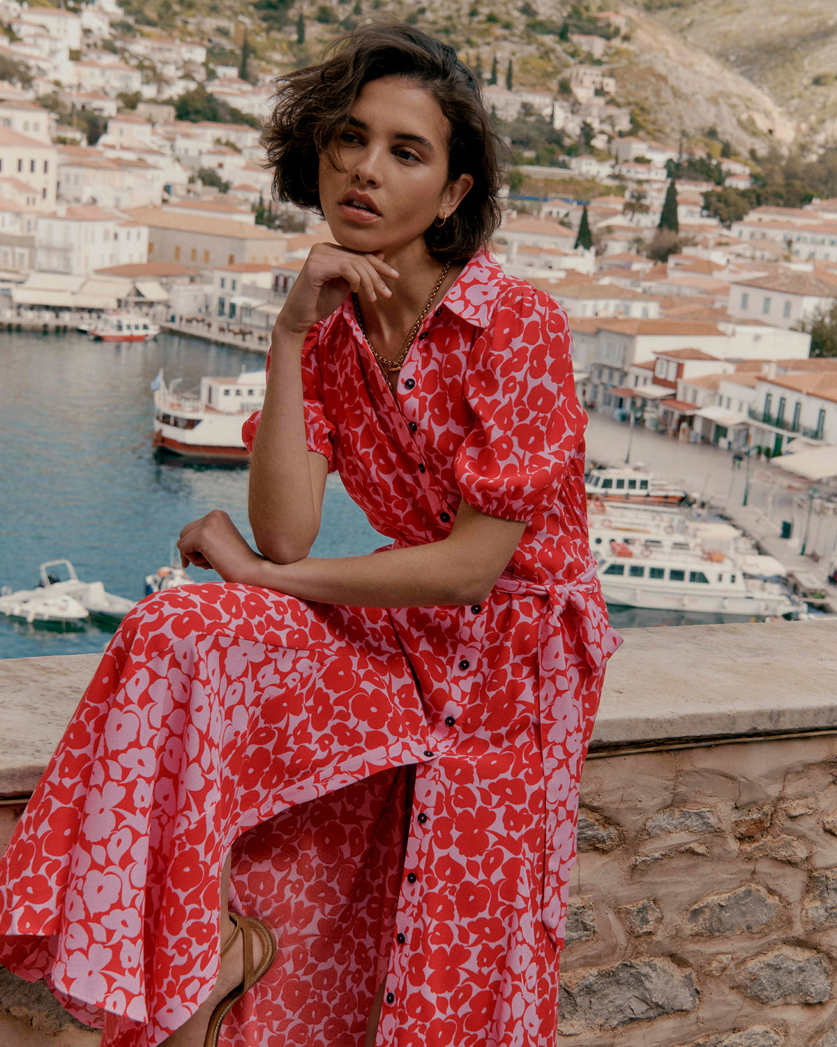 Woman in a red floral dress sitting by a harbor with a scenic town in the background